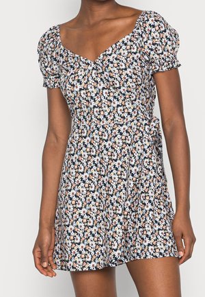 Kobieta ubrana w krótką, czarną sukienkę w kwiaty z wzorem białych i pomarańczowych stokrotek, z bufkami na krótkich rękawach i dekoltem w kształcie serca.