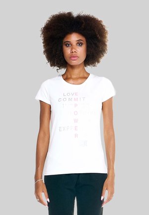 Femme aux cheveux bouclés portant un t-shirt blanc avec des mots verticaux pâles formant "EMPOWER" et un pantalon noir, debout devant un fond gris.