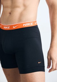 Black boxer briefs with a bright orange Nike logo waistband. Made of smooth fabric with a snug fit and contrasting small logo on the left leg.