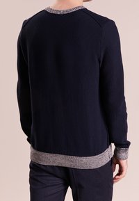 Homme portant un pull en maille bleu marine avec des poignets, un col et un ourlet côtelés gris, vu de dos sur un fond uni.