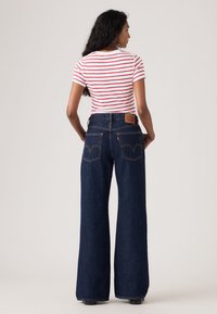 Naine seisab seljaga, kandes valget ja punase triibulise lühikese varrukaga särk ning kõrge vöökohaga laia säärtega tumedat sinist teksapüksid.