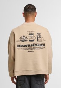 Sweatshirt oversize beige con illustrazioni nere stampate di cibi per la colazione e testo riguardante "Colazione per la Sbornia" sul retro.