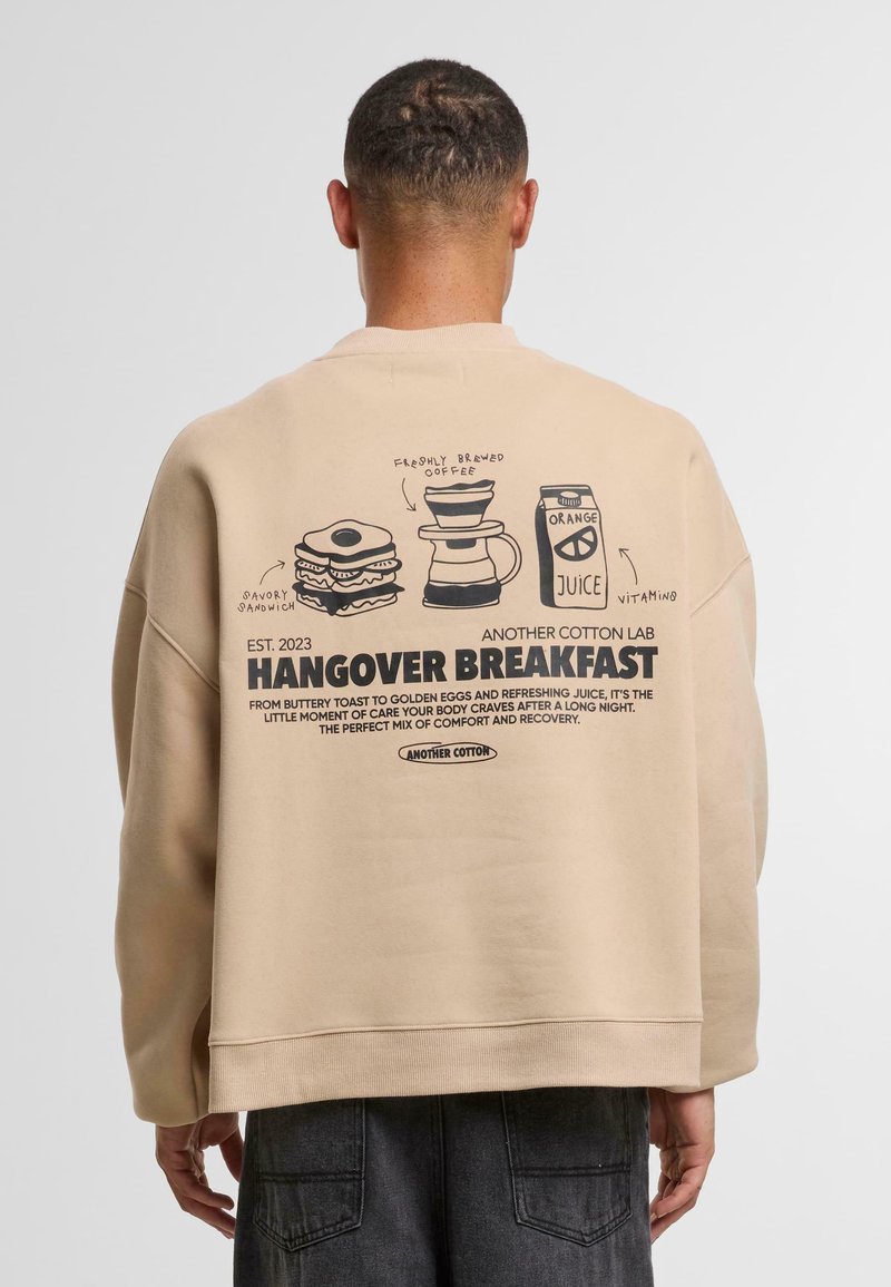 Sweatshirt oversize beige con illustrazioni nere stampate di cibi per la colazione e testo riguardante "Colazione per la Sbornia" sul retro.