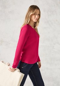 Långärmad topp med zigzag-mönster i klar rosa, matchad med mörkblå jeans. Personen håller en krämfärgad, fluffig väska.