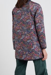 Kukkaaiheinen tikkitakki tummissa sävyissä, jossa on pyöreä kaula-aukko ja pitkät hihat, yhdistettynä samettisiin vihreisiin housuihin.