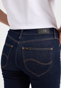 Persoon draagt donkerblauwe jeans van het merk Lee met zichtbare achterzakken, contrasterende stiksels en een wit ingestopt overhemd.