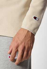 Fleece mikina světle béžové barvy s texturovanou tkaninou; obsahuje malou vyšitou nášivku na rukávu a šedou látku uvnitř.