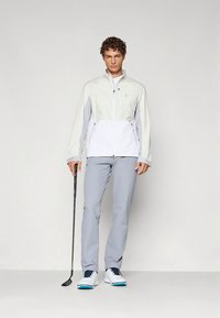Heren golfoutfit: lichtgrijze broek, witte en lichtgrijze jas van een stoffenmix. Effen schoenen met blauwe accenten. Golfclub in de hand.