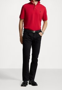 Červené polo tričko s límečkem a dvěma knoflíky, kombinované s černými džíny. Doplňky zahrnují černý opasek a stříbrné hodinky.