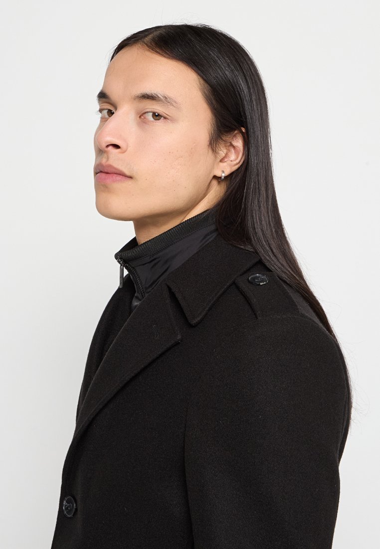 Schwarzer Wollmantel mit doppelter Knopfleiste und Schulterklappen. Darunter eine schwarze Jacke mit hohem Kragen und Reißverschlussdetails. Lange, glatte Haare.