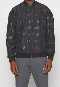 Svart bomberjacka med präglat "ADIDAS" logomönster, ribbade manschetter och nederkant, dragkedjestängning och en slät, glänsande yta.