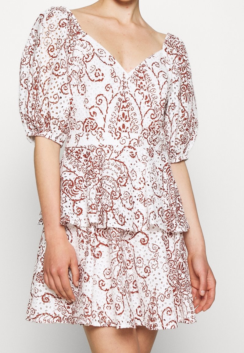 Femme portant une robe blanche avec des manches bouffantes et des motifs floraux bruns complexes en spirale, avec un tissu œillet et une jupe à volants.