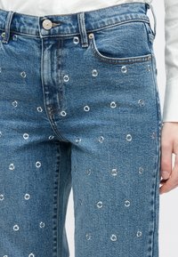 Mėlynos spalvos džinsai su sidabriniais akelėmis, pasklidusiais per audinį, ir tradiciniu penkių kišenių dizainu.