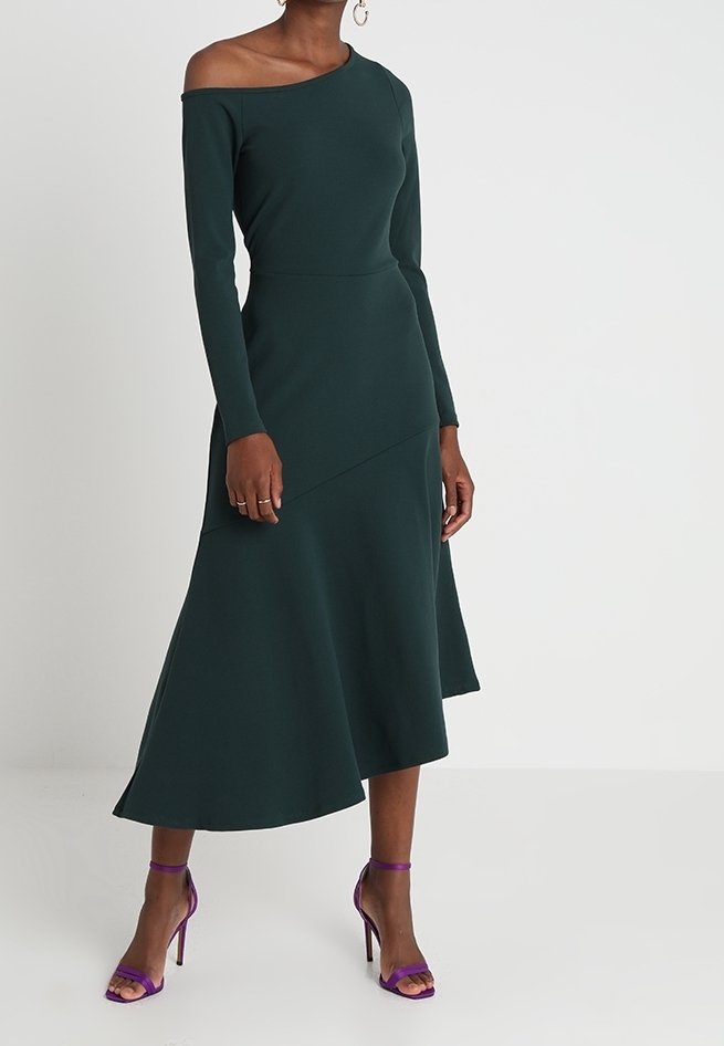 Robe midi vert foncé, épaules dénudées, manches longues et ourlet évasé. Le tissu semble lisse, accompagné de sandales à talons hauts violettes.