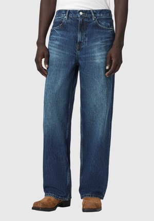 Mann trägt locker sitzende ausgewaschene blaue Jeans, weißes Oberteil und braune Wildlederstiefel, steht vor einem schlichten hellen Hintergrund.