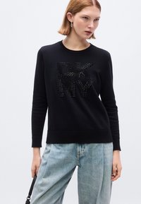 Felpa nera realizzata in tessuto morbido con logo DKNY impreziosito da strass. Maniche lunghe con polsini e orlo a costine. Jeans a lavaggio chiaro.