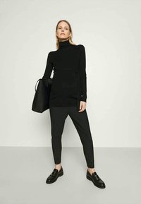 Črna turtleneck pulover s prilegajočo silhueto, v kombinaciji z črno-belimi prugastimi hlačami. Dodatki vključujejo veliko črno torbo in loafer čevlje.