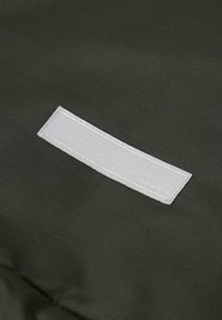 Tissu vert foncé avec une pièce rectangulaire horizontale gris clair cousue, affichant un texte subtilement embossé dans le coin inférieur droit.