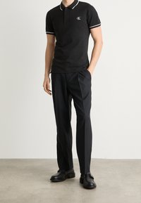 Černé polo tričko s bílými akcenty, černé kalhoty na míru a černé boty. Obsahuje malé logo na tričku a je požadován oblek na míru.