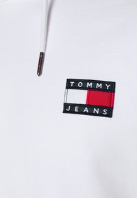 White garment with a rectangular stitched black patch showing a flag with white and red sections and "TOMMY JEANS" text above and below.