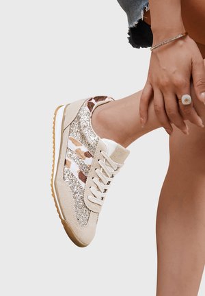 Pied chaussé d'une basket beige ornée de paillettes et de rayures à motif animalier, reposant sur le genou opposé ; main avec bague et bracelet tenant doucement la cheville.