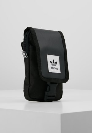 Bolsita de tela negra con cierre de solapa, presenta un parche con el logo de Adidas y una correa lateral; diseñada para almacenamiento compacto y portabilidad.