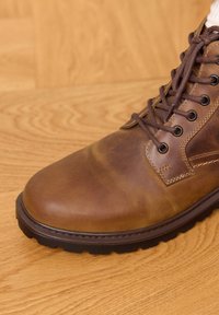 Brauner Leder-Schnürstiefel mit gesteppten Details und einer robusten schwarzen Gummisohle auf einem Holzboden.