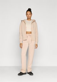 Ανοιχτό μπεζ αθλητικό σύνολο που περιλαμβάνει φούτερ με φερμουάρ, κροπ τοπ και φόρμες με κορδόνι. Τα παπούτσια είναι μαύρα με υφή σχεδίου.