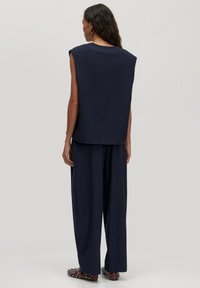 Vrouw met lang donker haar draagt een mouwloze marineblauwe top en een wijdvallende marineblauwe broek, gecombineerd met gestalde bordeauxrode schoenen, staand tegen een effen achtergrond.