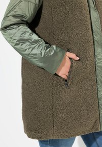 Grön textiljacka med en plyschig, strukturerad fleecekropp och släta, reflekterande ärmar. En dragkedjeficka på sidan ger praktisk detalj.