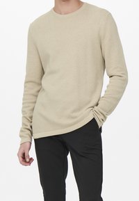 Beżowy, teksturowany sweter z długim rękawem, z okrągłym dekoltem, o luźnym kroju i detalem w pasie, zestawiony z czarnymi spodniami.