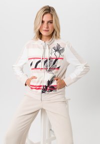 Lichtbeige ritsjack met bloemenprint in zwarte, rode en witte strepen. Heeft een hoge kraag en zijzakken. Gladde stoftextuur.