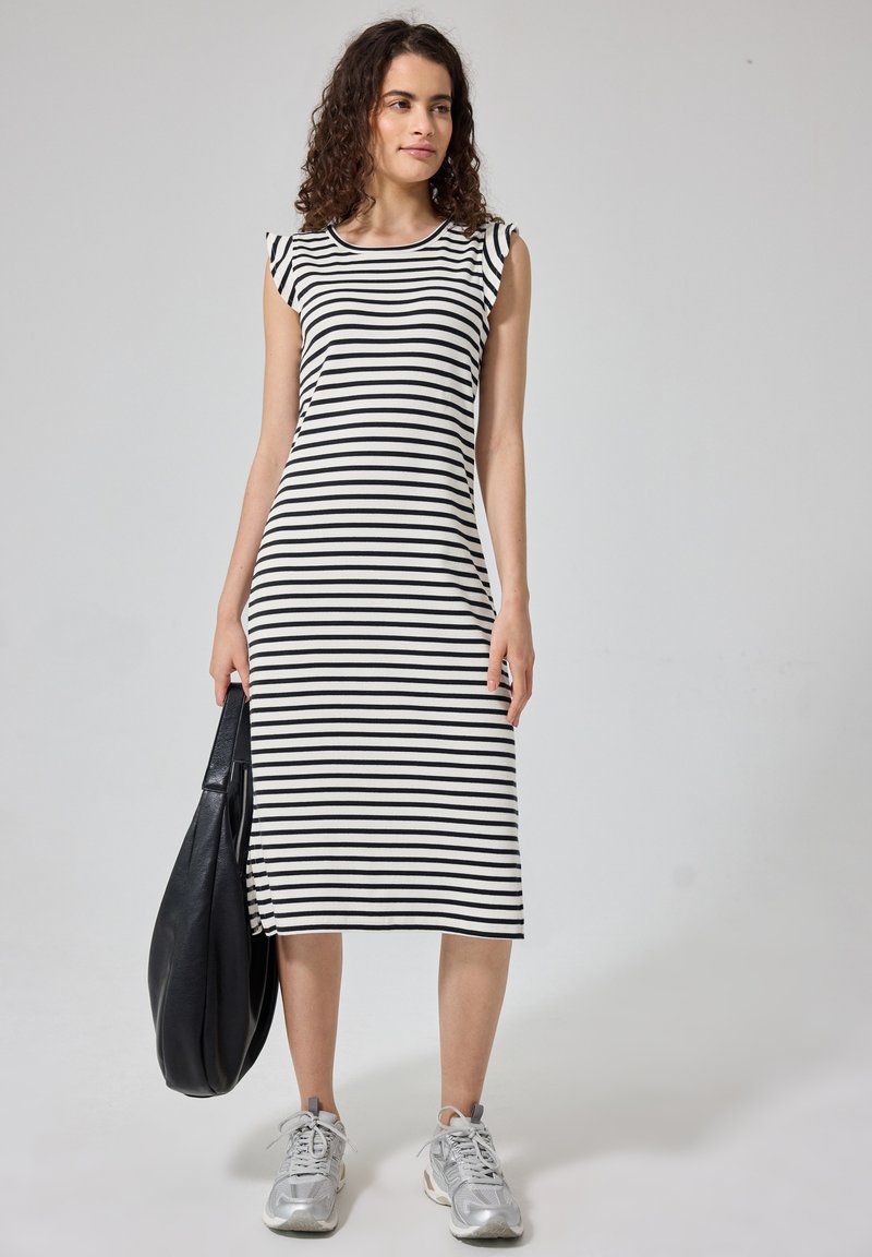 Jeune femme portant une robe sans manches rayée noir et blanc, tenant un grand sac noir avec des baskets argentées sur un fond uni.