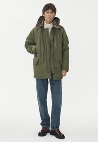 Olivnozeleni podloženi plašč z ovratnikom iz sive tkanine, dvema sprednjima žepoma in sprednjim zadrgo, oblečen čez kremni pulover s turtleneck ovratnikom in modre kavbojke.