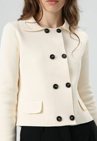 Veste croisée couleur crème en tissu texturé, avec des boutons noirs, un col cranté et deux poches avant.