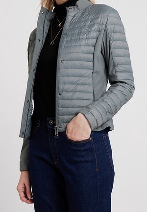 Femme portant une veste matelassée gris clair sur un col roulé noir, associée à un jean bleu foncé, se tenant avec une main dans la poche.