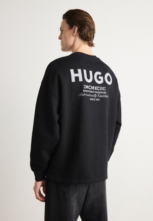 Mustan kapuutsiga särk, mille seljal on suur valge "HUGO" kiri, varrukate otsad on soonilised ja kangas on tekstuuriga. Kombineeritud tumedate teksadega.