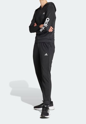 Person stående med armene krydsede iført sort Adidas træningsdragt med hvidt logo på ærmet, bukserne og sorte Adidas-sneakers.