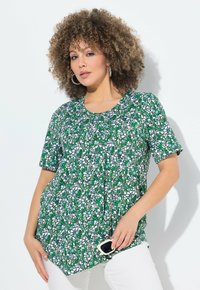 Top corto a maniche corte verde con motivo che presenta un collo rotondo, realizzato in tessuto leggero con un mix di forme bianche e verdi. Pantaloni bianchi e occhiali da sole.