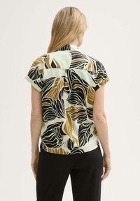 Blusa a maniche corte di un leggero colore menta con un motivo di foglie nere e dorate. Il retro presenta un yoke con una vestibilità rilassata e un orlo leggermente curvo.