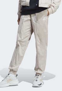 Beige joggerbyxor med resår i ärmsluten och sidofickor, med vita broderade logotypdetaljer. Tillverkade av ett mjukt tyg med en avslappnad passform.