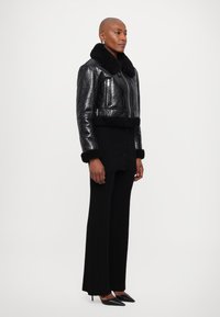 Jachetă din piele neagră cu un finisaj lucios și guler de blană, asociată cu pantaloni negri cu model în dungi și tocuri negre ascuțite. Design simplu cu accente minime.