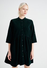 Robe en velours côtelé vert foncé avec un col arrondi, devant boutonné, manches trois-quarts et une taille froncée pour une silhouette évasée.
