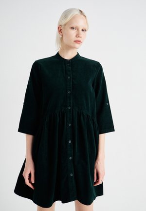 Robe en velours côtelé vert foncé avec un col arrondi, devant boutonné, manches trois-quarts et une taille froncée pour une silhouette évasée.