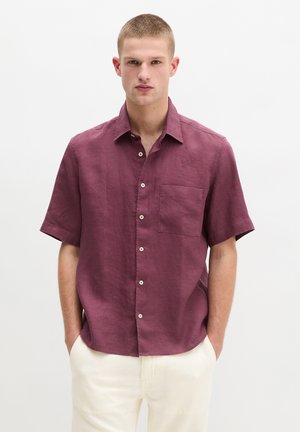 Junger Mann mit kurzen Haaren, der ein bordeauxrotes Hemd mit kurzen Ärmeln und helle Hosen trägt, die Hände in den Taschen.