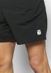 Lyhyt mustat shortsit kevyestä kankaasta, joissa on pieni valkoinen logo-laatta, jossa on kasvokuvio helman lähellä. Sileä rakenne, polvipituiset.