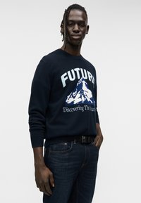 Πλεκτό πουλόβερ σε ναυτικό μπλε χρώμα με τη λέξη "ΜΕΛΛΟΝ" και γραφικό βουνό. Συνδυάζεται με σκούρο μπλε τζιν, παρουσιάζοντας άνετη γραμμή.