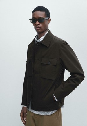 Unge mand iført mørke solbriller, mørk knappet jakke med store lommer, stribet skjorte og beige bukser mod en ensartet lys baggrund.