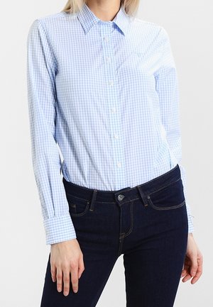 Femme portant une chemise longue à boutons à carreaux bleu clair et blanc et un jean bleu foncé, debout avec une main le long du corps.