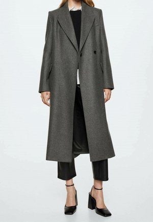 Kobieta w długim szarym dwurzędowym płaszczu, na czarnych skróconych spodniach i czarnych butach na wysokim obcasie z paskami wokół kostek.